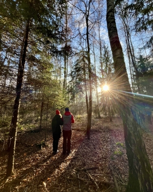 Dwie osoby stojące koło siebie w gęstym lesie patrzące na promienie słońca przenikające przez drzewa (c) Łukasz Derkacz